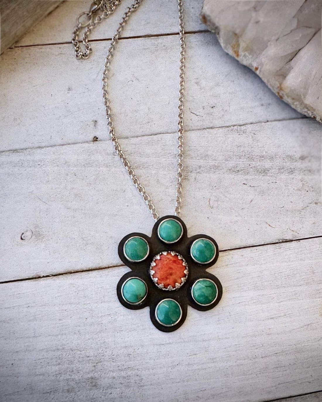A handmade necklace shaped like a flower with a round orange coral stone center and turquoise petals.