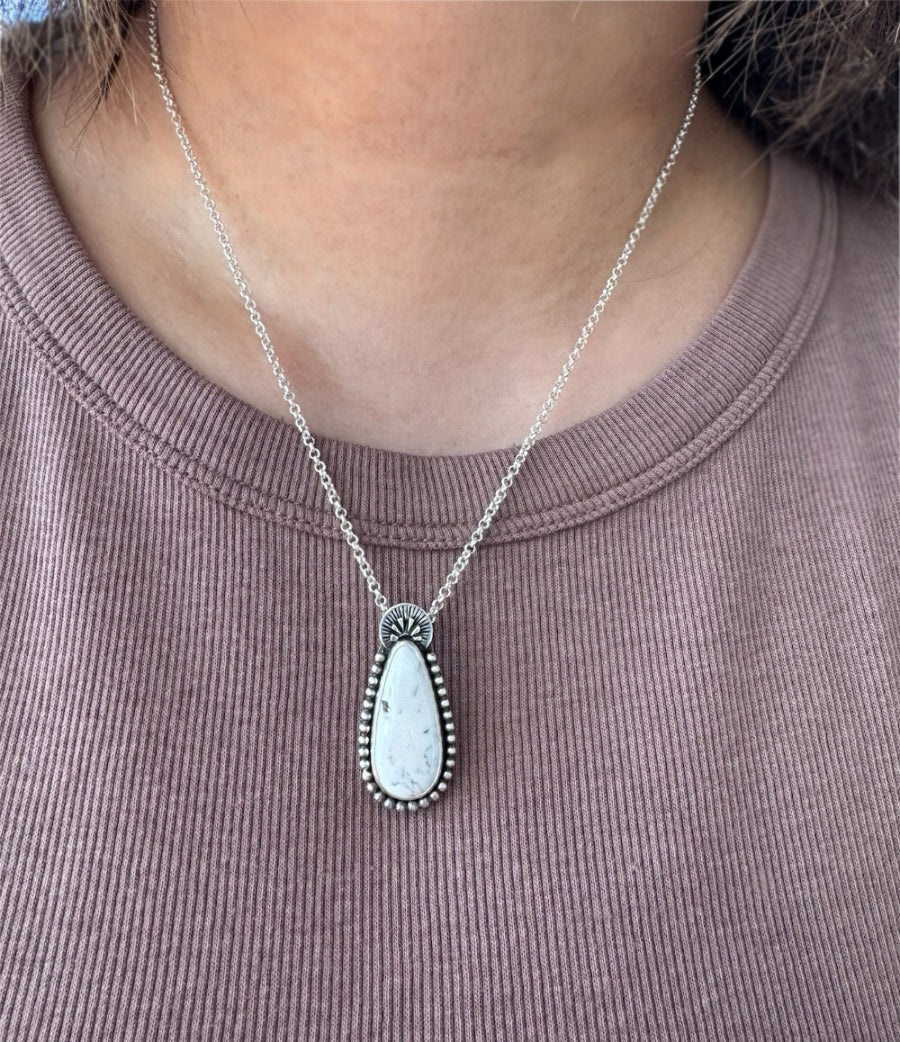 A handmade sterling silver necklace with a white buffalo teardrop shaped stone as the focal point, with silver balls wrapped around it and a handstamped arc at the top shown worn around the neck.