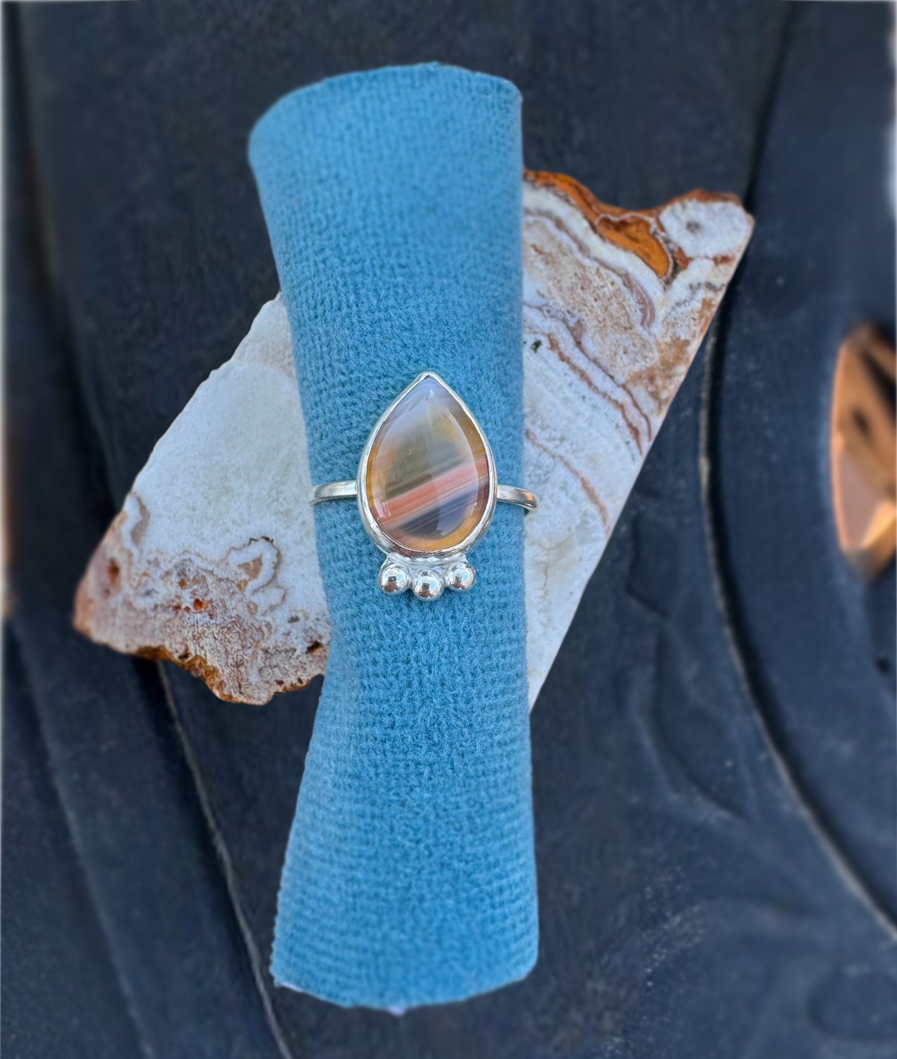 Handmade sterling silver ring with a teardrop shaped Botswana Agate gemstone and 3 silver ball embellishments on the bottom on a blue fabric and dark background.