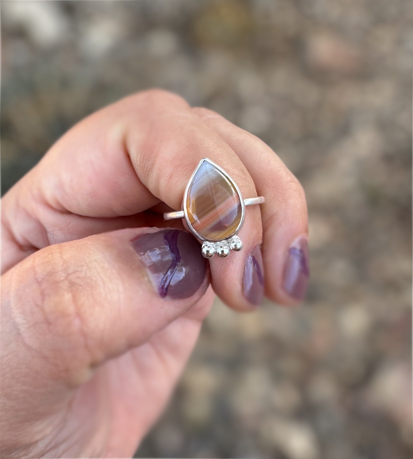 Teardrop-shaped Botswana agate ring with a silver band held between fingers against a blurred background
