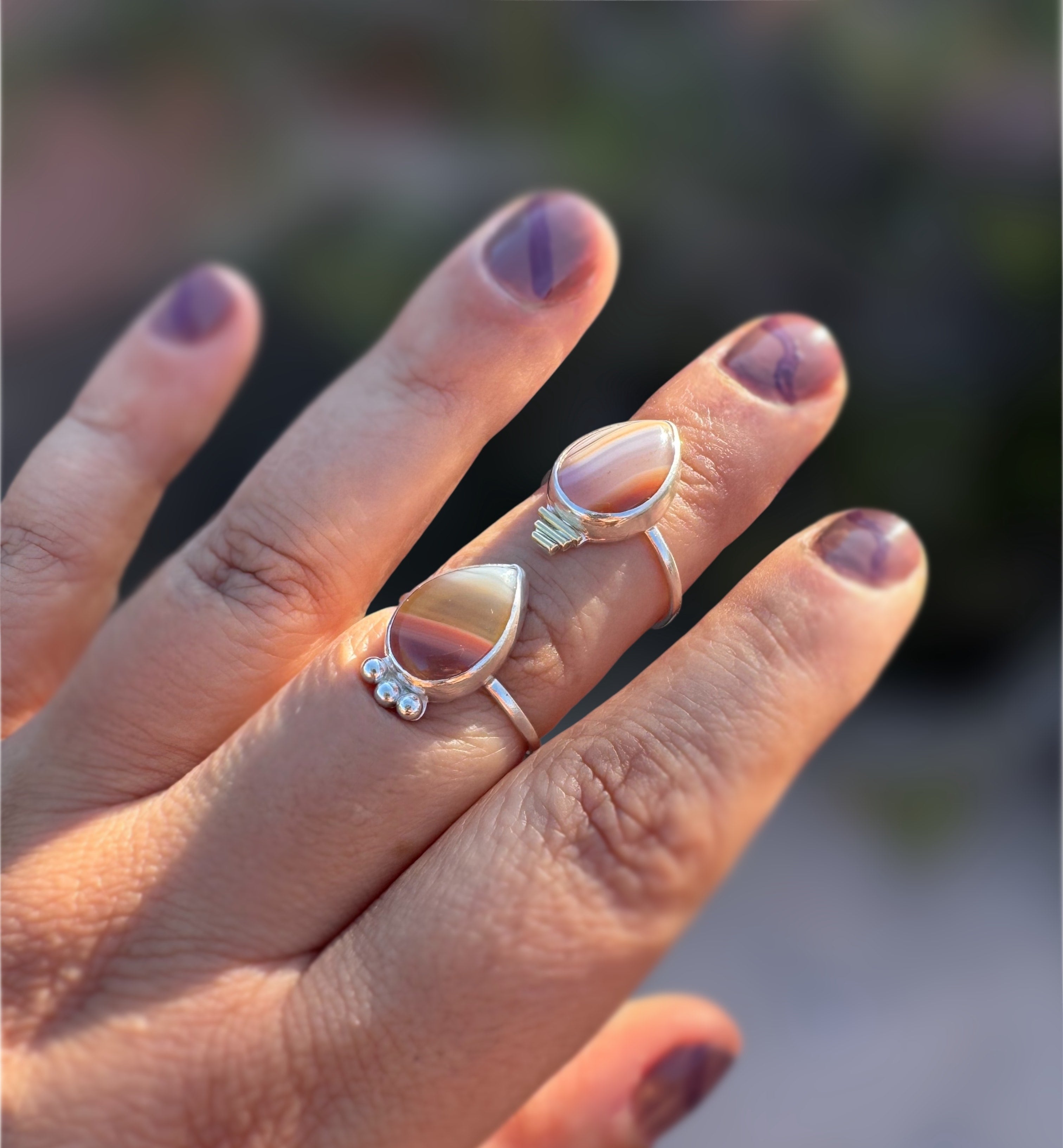 Hand wearing two sterling silver rings with peach and pink colored teardrop-shaped Botswana Agate gemstones against a blurred natural background