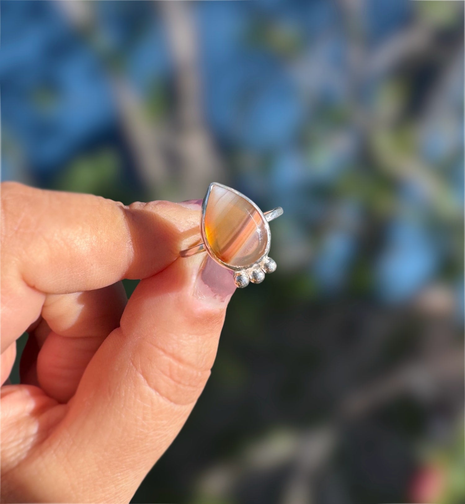 Teardrop-shaped Botswana agate ring with a silver band held between fingers against a blurred background