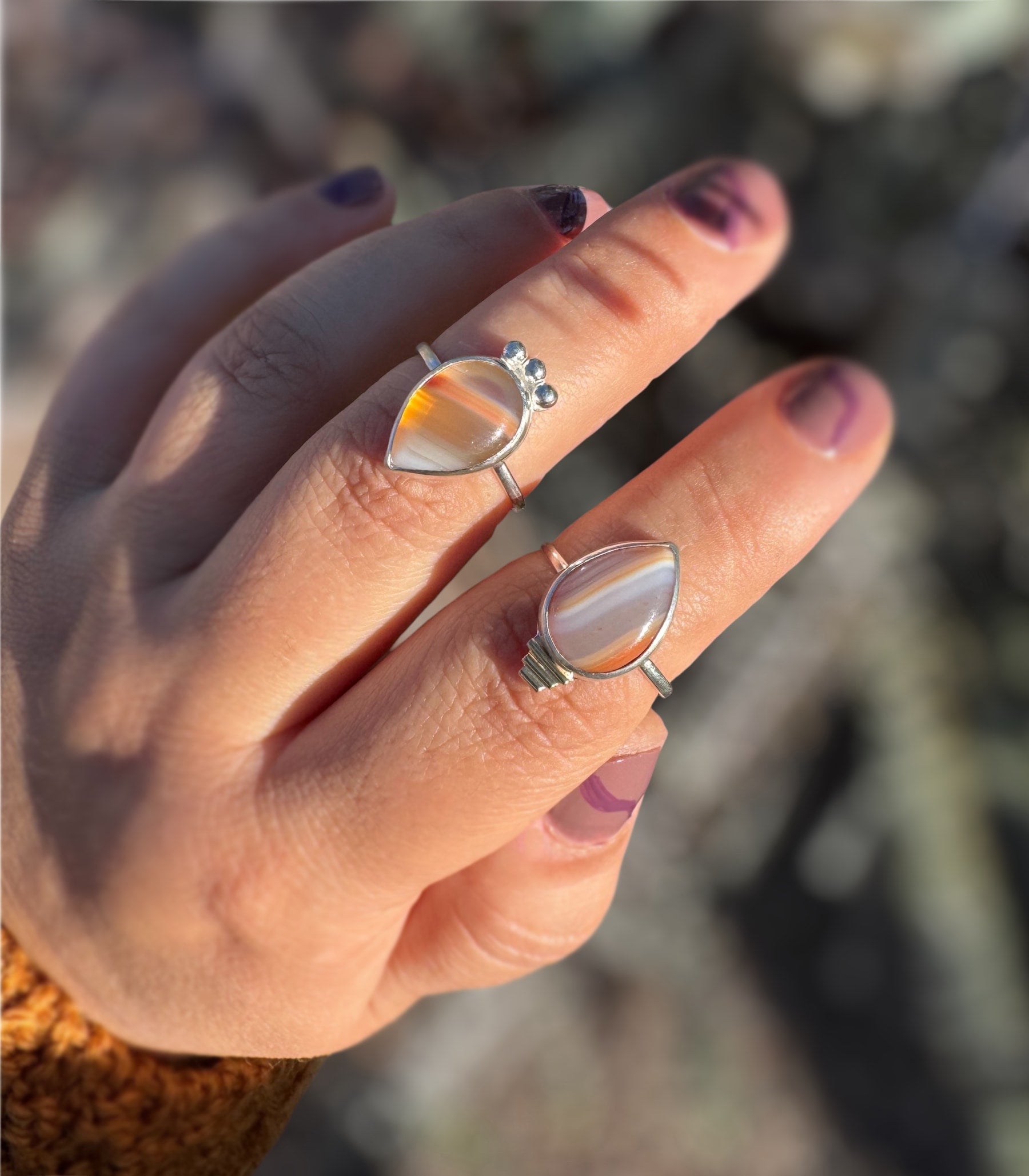 Hand wearing two sterling silver rings with teardrop-shaped Botswana Agate gemstones against a blurred natural background