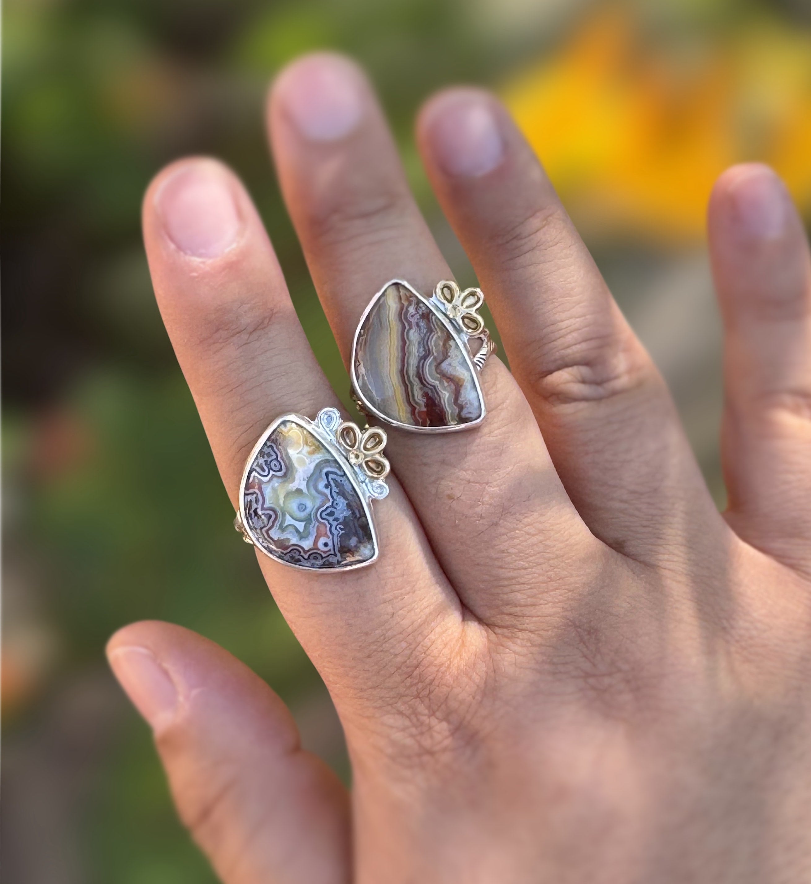Two silver rings with crazy lace agate stones are shown on a person's fingers, with an outdoor background.