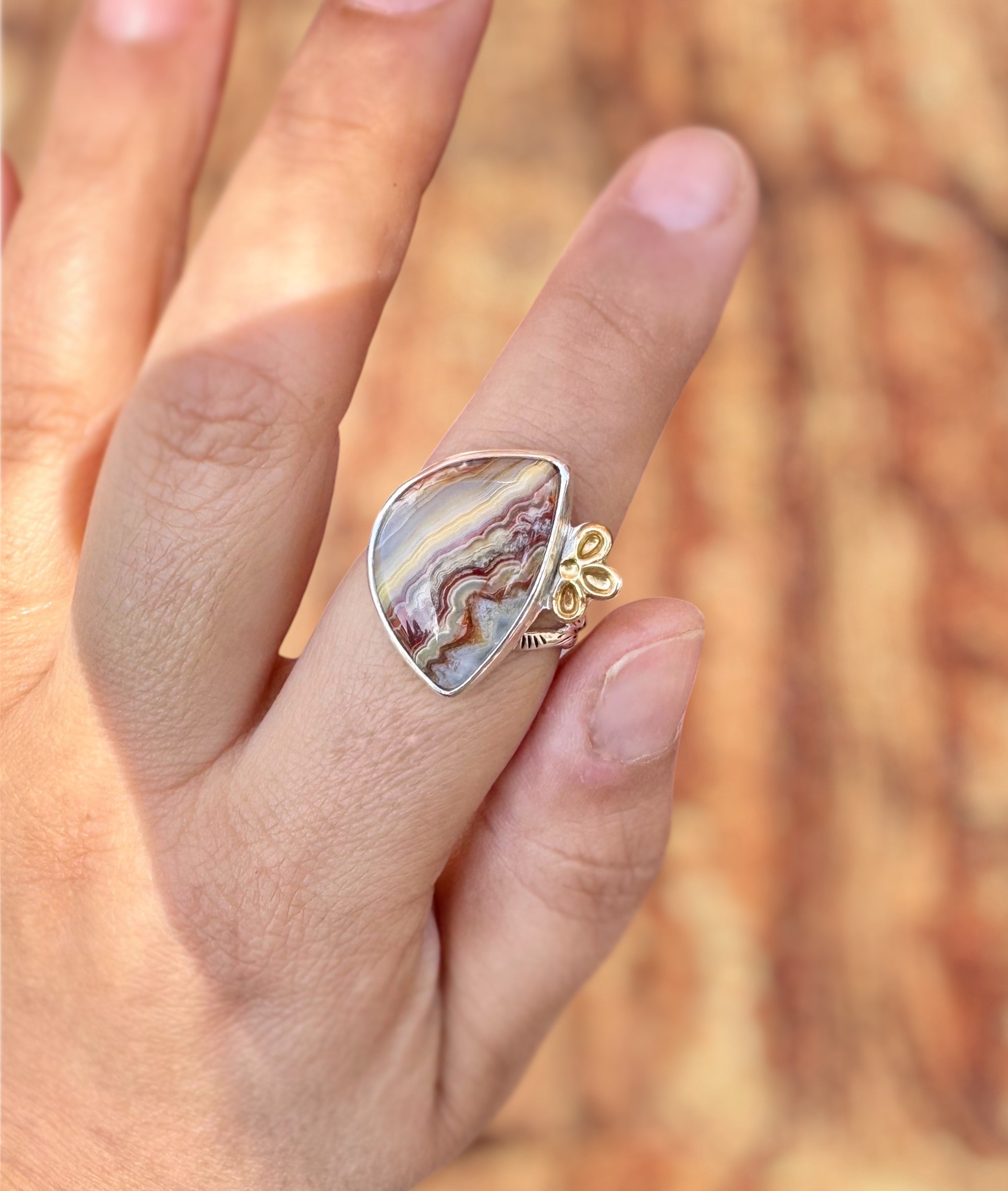 A close-up of a hand wearing a unique ring featuring a large, irregular teardrop-shaped agate stone with natural brown and cream banding, set in a silver bezel with a small gold floral detail on the side, against a warm, blurred wood background.