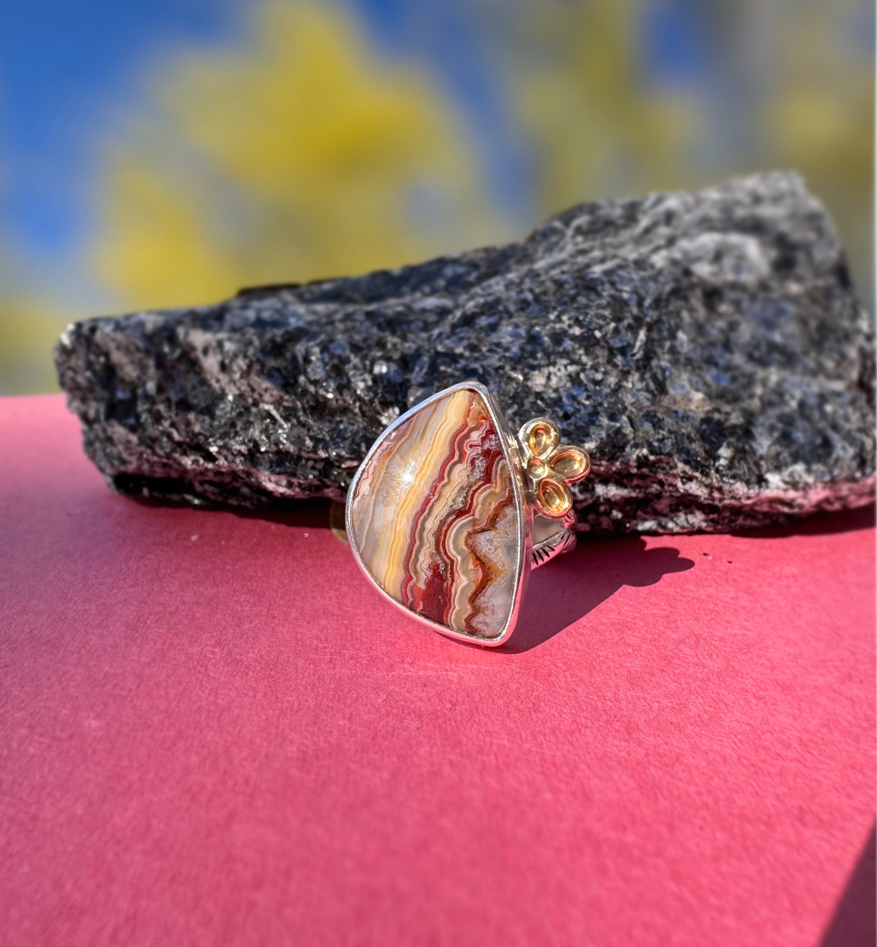 A silver ring with a crazy lace agate stone featuring red, orange, and cream banded patterns, displayed on a pink surface with a dark rock and a blurred outdoor background.