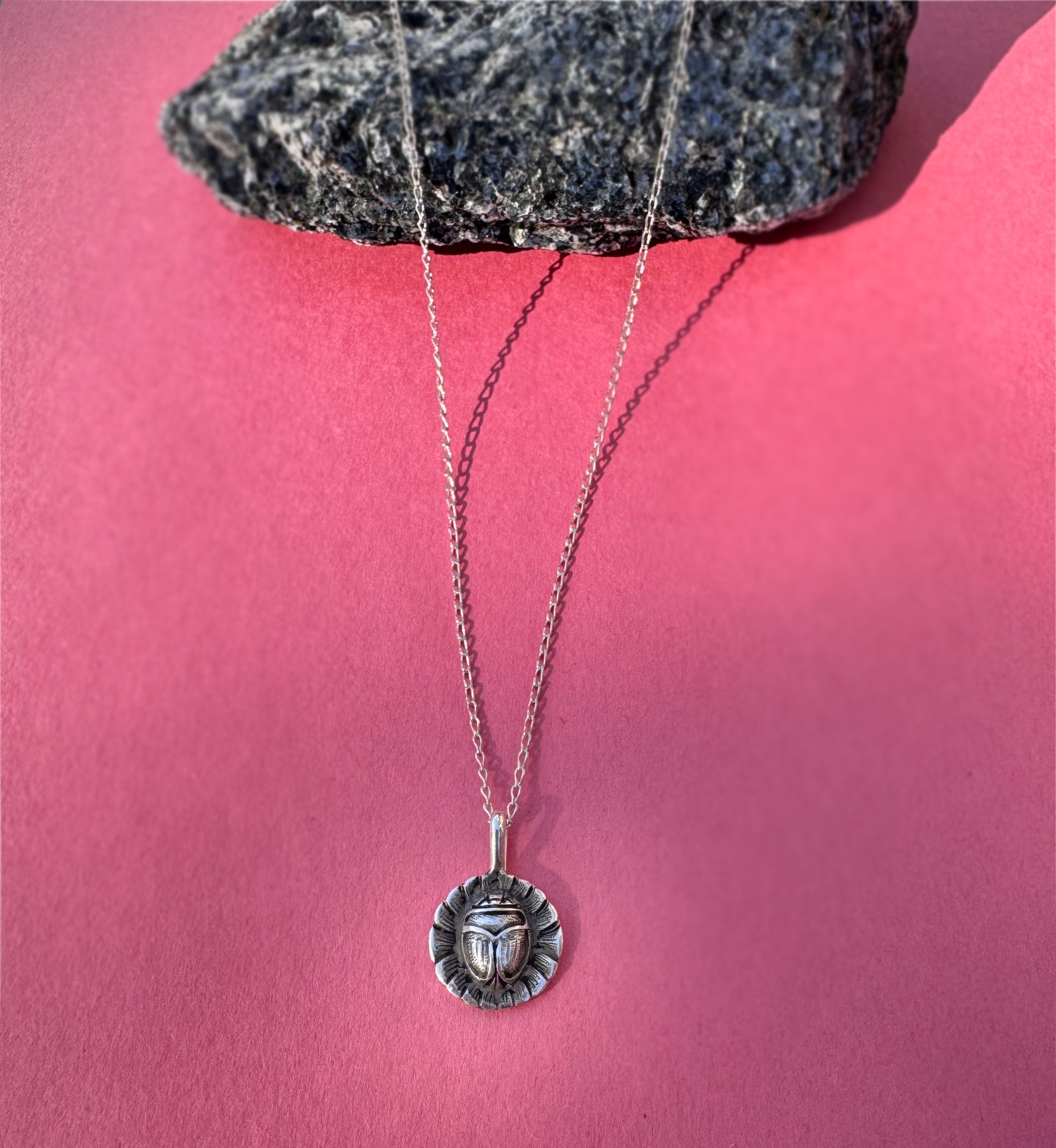A silver pendant necklace with a circular medallion featuring a stylized scarab on a flower, displayed on a pink background with a dark gray rock prop at the top.
