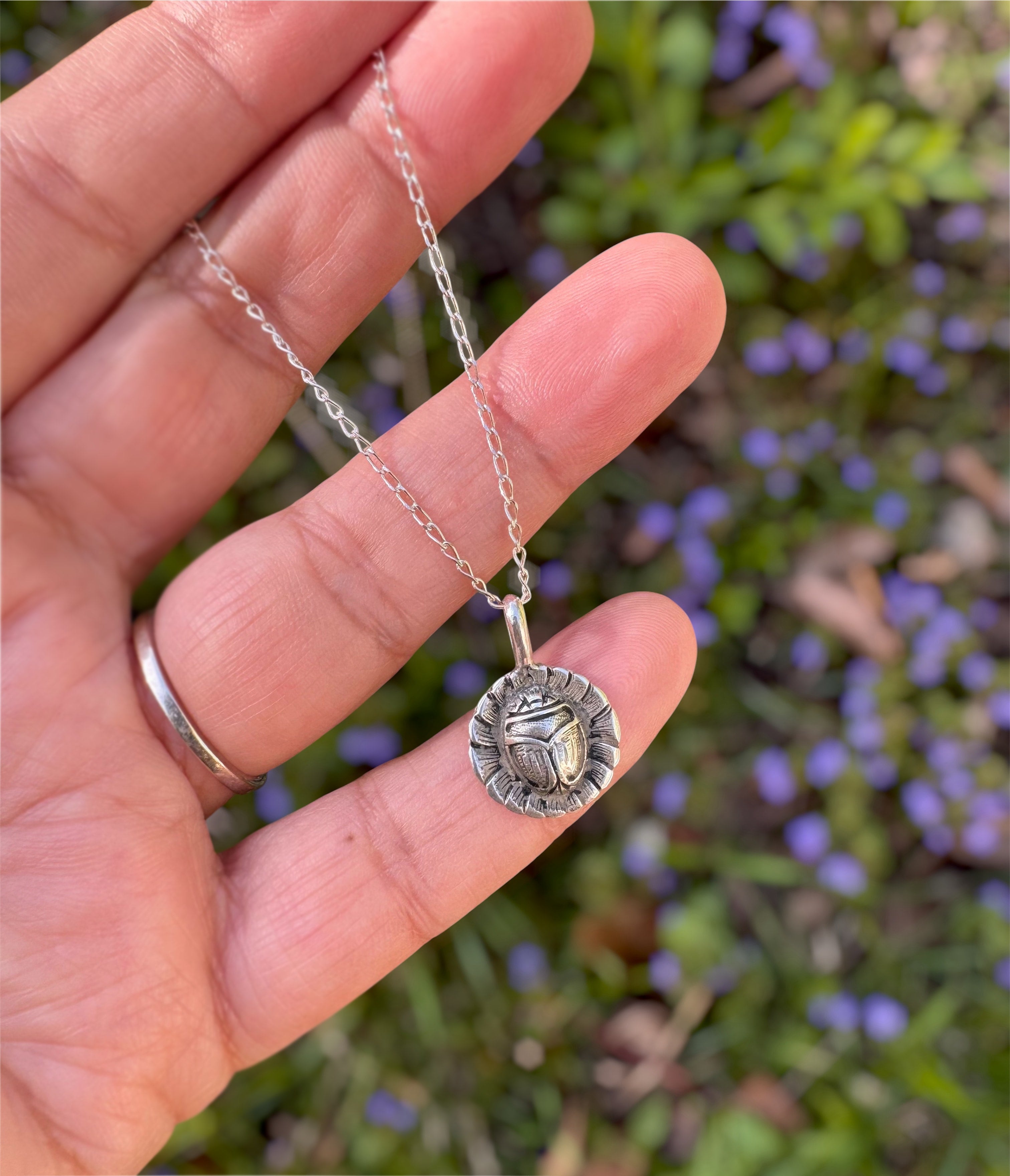A hand holding a silver chain necklace with a circular pendant featuring a scarab beetle design, set against a blurred background of green foliage and purple flowers.