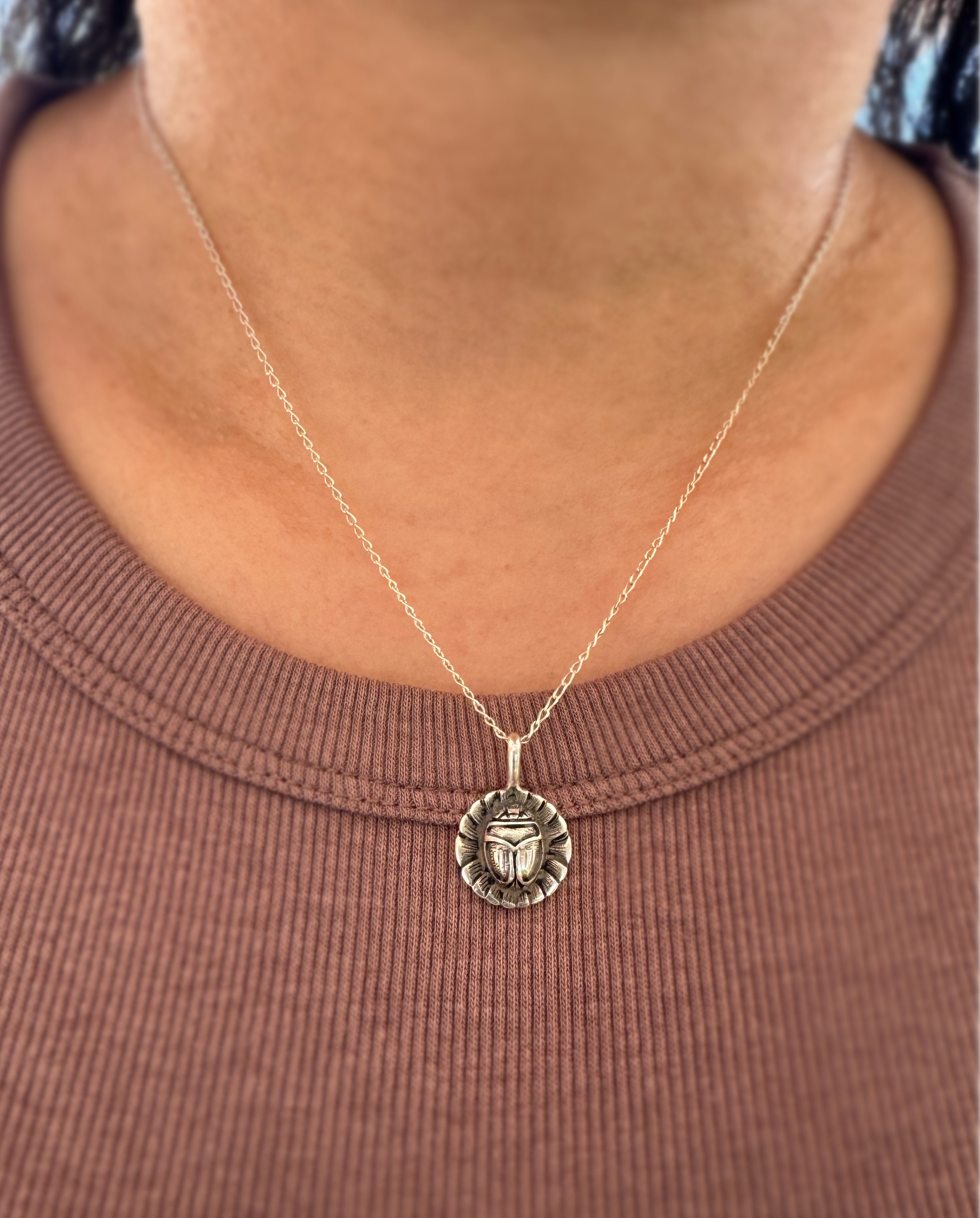 A close-up of a person wearing a silver chain necklace with a circular pendant featuring a scarab beetle design on a flower, styled with a brown ribbed crewneck shirt.