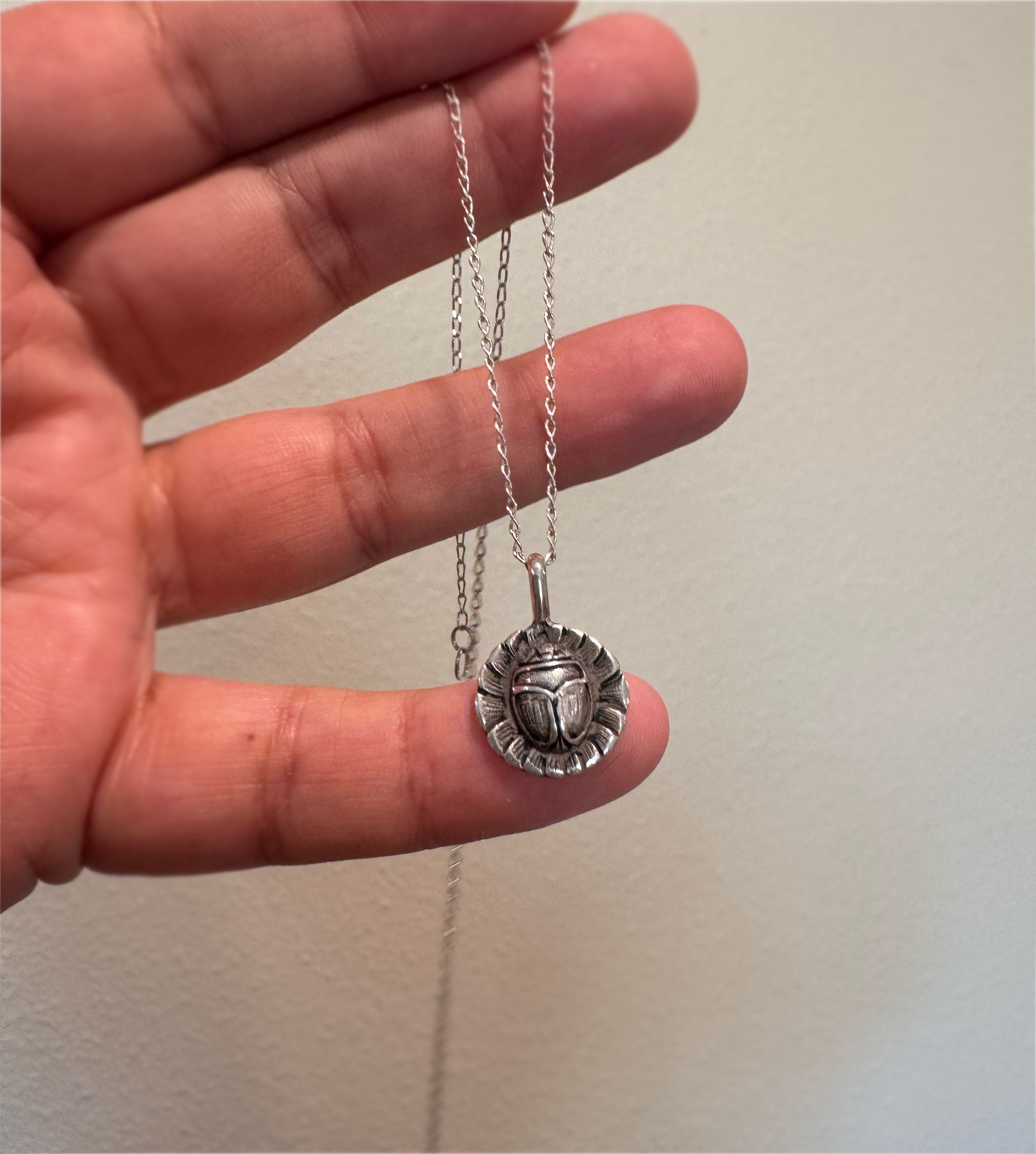 A sterling silver chain necklace with a circular scarab beetle pendant is held in a person's hand against a plain light beige wall.